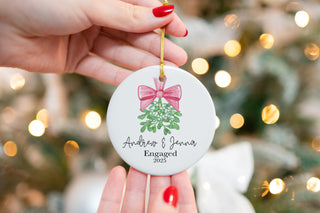 Custom ceramic ornament featuring mistletoe with a pink bow, personalized with couple’s names “Andrew & Jenna” and year 2025 — engagement keepsake.