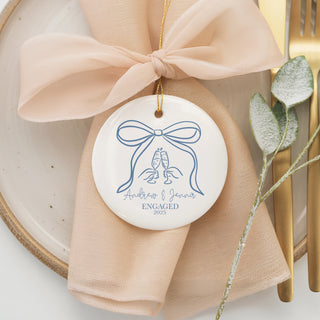 Custom ceramic ornament featuring a large blue bow with clinking champagne glasses, personalized with couple’s names “Andrew & Jenna” and year 2025 — engagement keepsake.