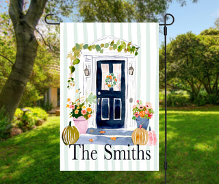Personalized fall garden flag featuring a watercolor navy front door with a floral wreath, pumpkins, flower pots, and a custom family name on a sage striped background.