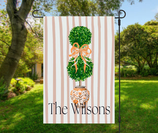 Personalized topiary garden flag featuring a boxwood topiary with a peach bow in a chinoiserie planter and a custom family name on a blush striped background.